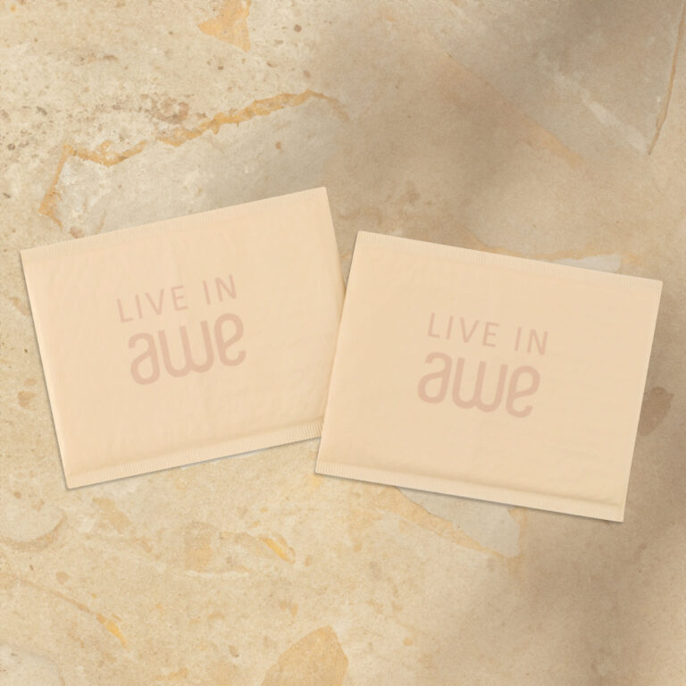 Two beige packets are placed on a textured beige surface. Both packets have the text LIVE IN awe printed on them. The overall tone is neutral and minimalist.