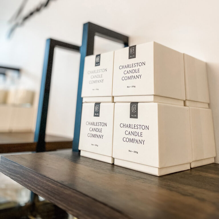 Stack of white boxes labeled Charleston Candle Company displayed on a dark wooden shelf. Geometric black frames are in the background against a white wall. The scene is stylishly minimalistic and tidy.