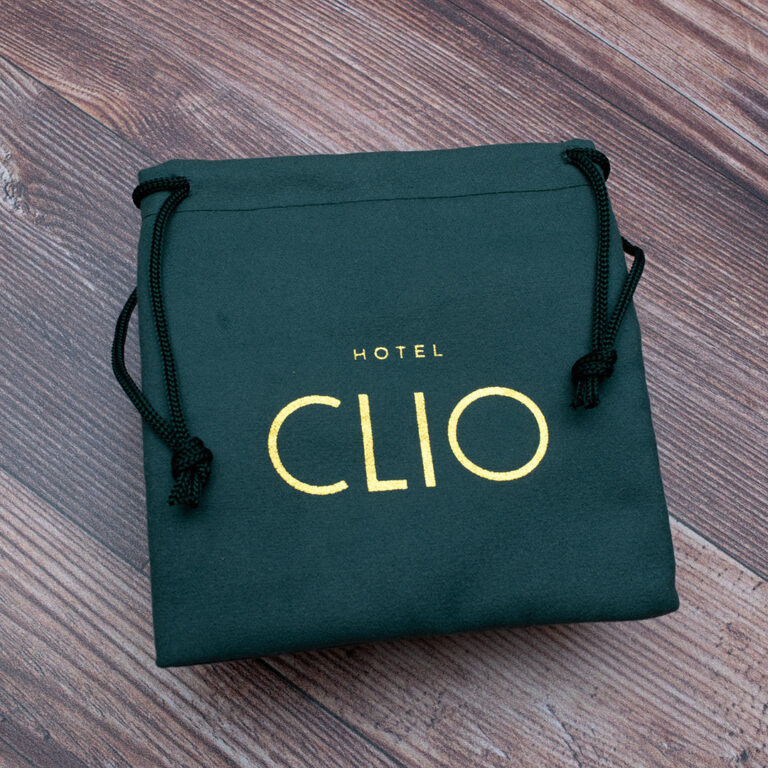 A green drawstring bag with Hotel Clio printed in gold sits on a wooden floor.