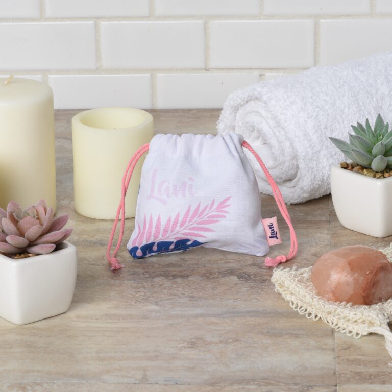 A small drawstring pouch with pink lettering and leaf design sits on a spa-themed surface. Surrounding it are candles, potted succulents, a folded white towel, a brown soap bar, and a loofah, all set on a light-colored tiled background.
