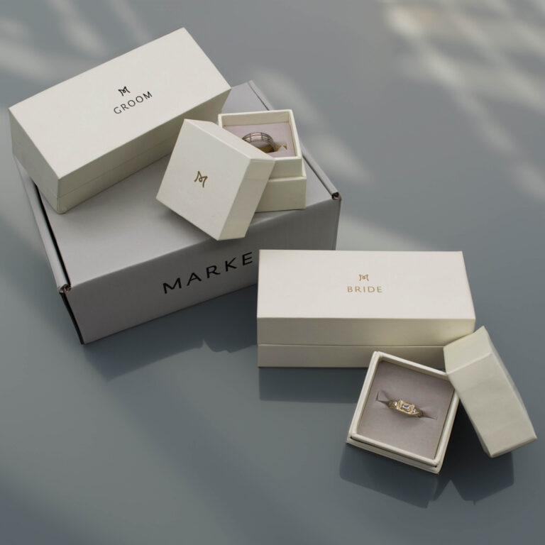 A set of elegant jewelry boxes labeled BRIDE and GROOM are displayed. Each box contains rings. The boxes are stacked on another larger box labeled MARKE, arranged on a glossy gray surface.
