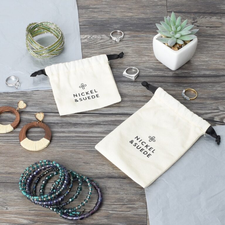 A flat lay of jewelry on a wooden surface, including a succulent in a white pot, colorful beaded bracelets, silver rings, wooden earrings, and two drawstring bags labeled Nickel & Suede.