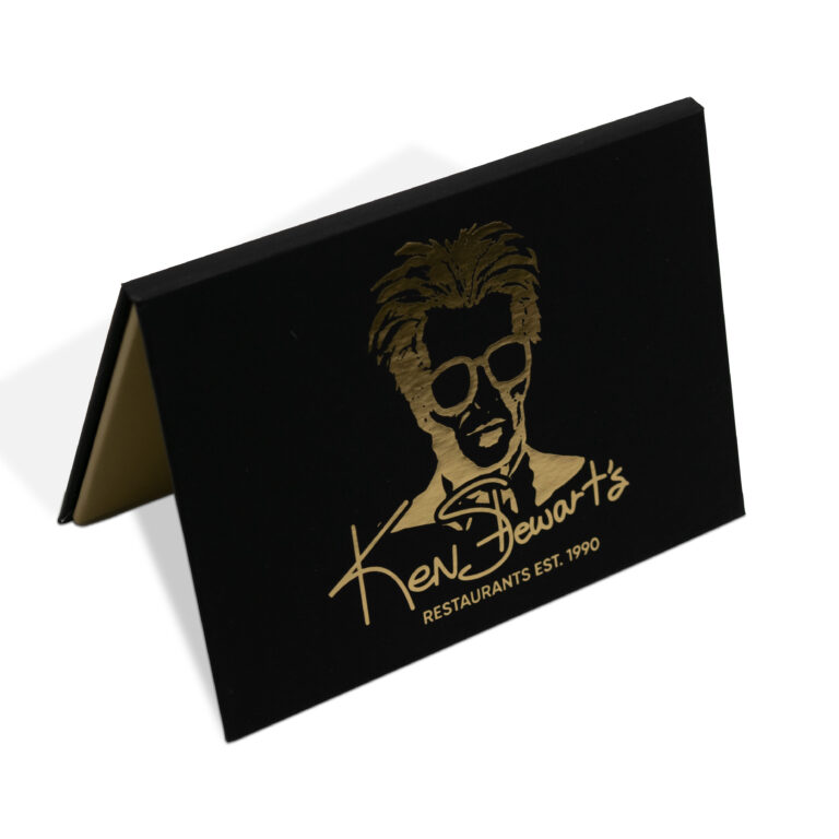 A black tent card featuring a gold illustration of a person wearing sunglasses and styled hair, with the text Ken Stewarts Restaurants Est. 1990 in gold lettering below the illustration.