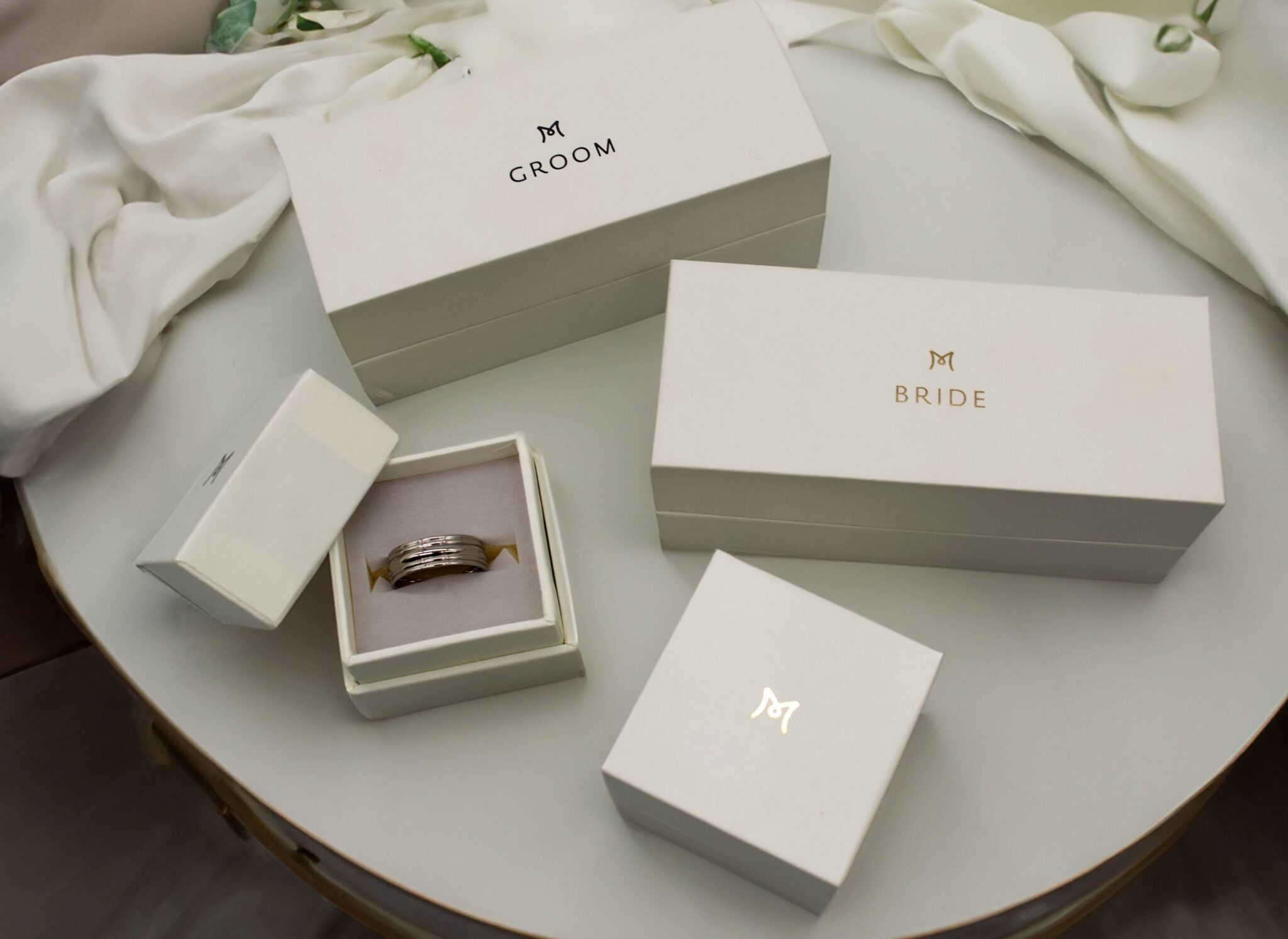 A table with elegant white ring boxes labeled Groom and Bride. One smaller box is open, revealing a set of rings. The boxes feature a simple monogram logo, and they are placed on a smooth, round surface.