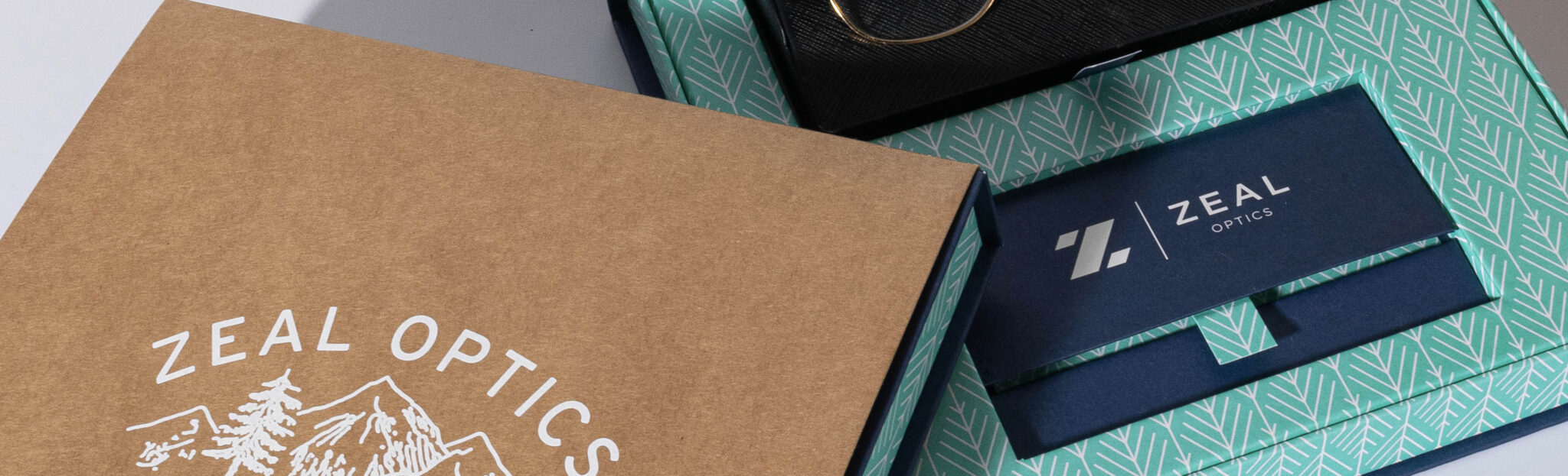 Boxes with Zeal Optics branding are partially open, revealing sleek eyewear on a patterned green-and-white interior. The outer lid is brown with mountain graphics and the distinctive Zeal Optics logo.